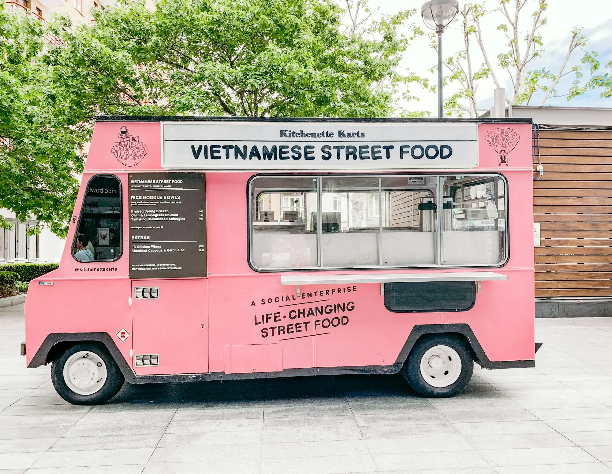 Pink converted street food van with a menu board and serving hatch, parked on a paved plaza under trees