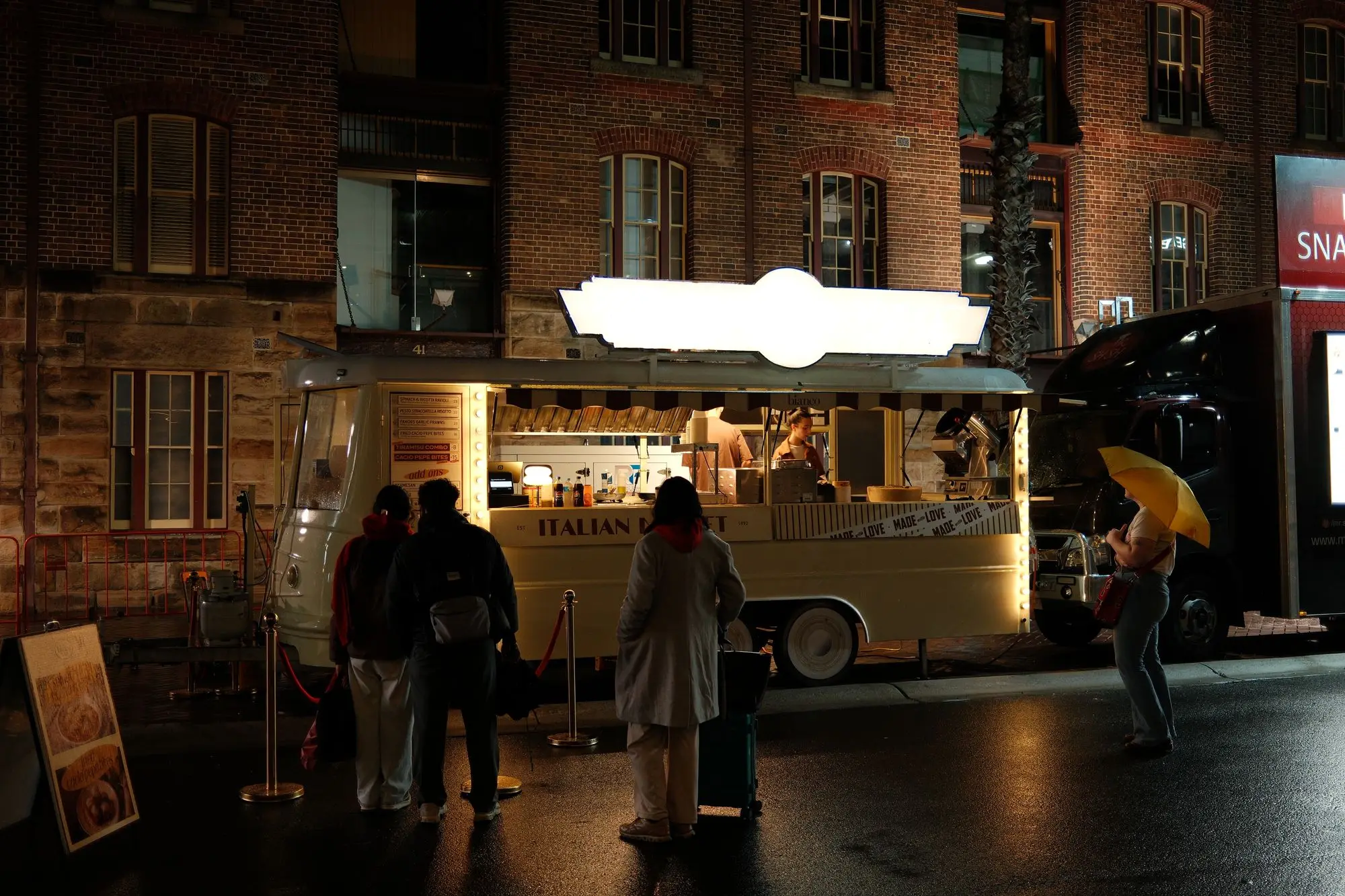 Customers queue at a food truck's hatch on a wet night in Sydney's The Rocks while a passer-by shelters under a yellow umbrella